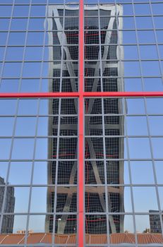 One of the twin Kio towers reflected on another one.  The towers are office buildings in Madrid, Spain .They were designed by the American architects Philip Johnson and John Burgee, commissioned by the Kuwait Investment Office (hence their initial name "Torres KIO" or "KIO Towers"). Each building is 115 metres tall with an inclination of 15 degrees, making them the first inclined skyscrapers in the world.