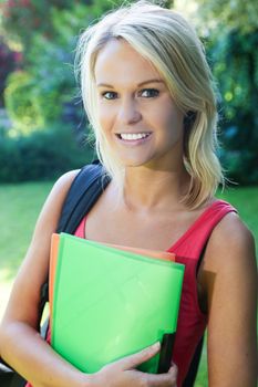 Pretty smiling blonde student with files in hand standing outdoors