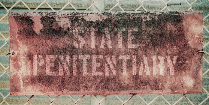 Retro Filtered Photo Of Rusty Grungy Old Penitentiary Prison Sign On Chain Link Fence