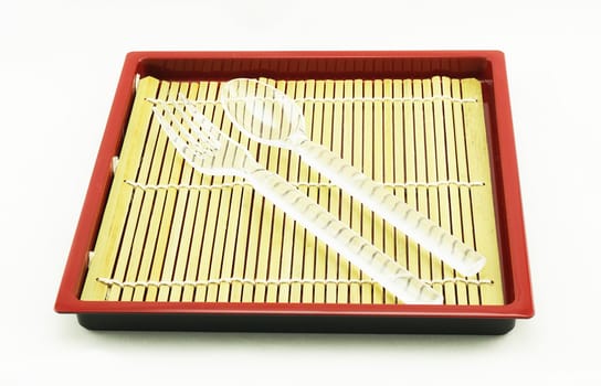 Spoon and fork are white color, placed on bamboo pad in wooden tray.                               