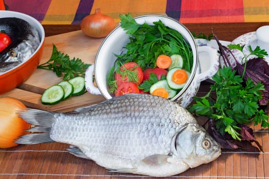 On a table on a chopping board there are a fish, cucumbers, tomatoes, onions, spices and parsley greens.