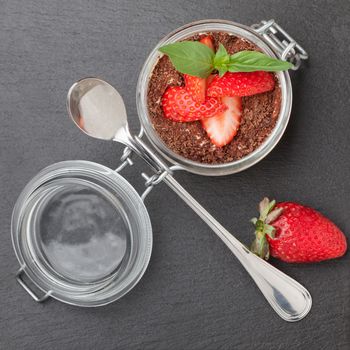 Delicious traditional italian cream dessert pannacotta with grated chocolate, strawberry, and basil served in jar with cocktail spoon on black stone background