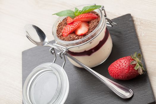 Delicious traditional italian cream dessert pannacotta with grated chocolate, strawberry, and basil served in jar with cocktail spoon on black stone background