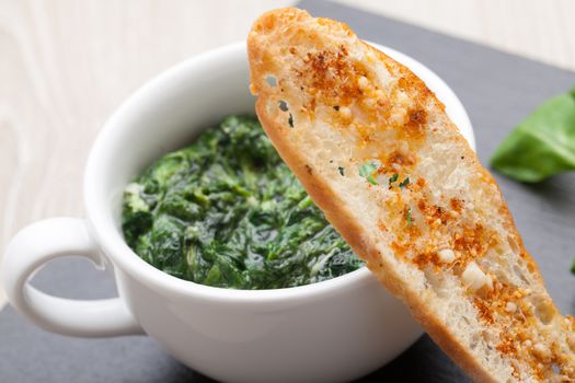 Appetizer sauteed garlic spinach dish in cup, baked bread slice  with  melted cheese served on black stone