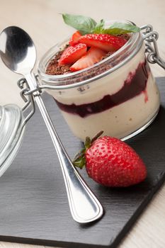 Delicious traditional italian cream dessert pannacotta with grated chocolate, strawberry, and basil served in jar with cocktail spoon on black stone background