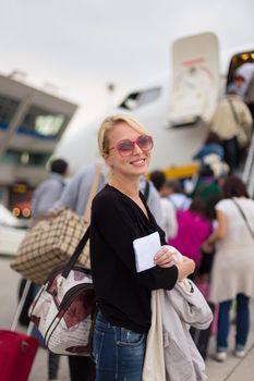 Cheerful woman holding carry on luggage queuing to board the commercial airplane.