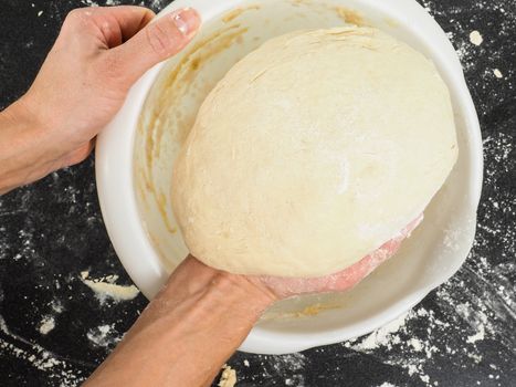 Person lifting a proven dough out from a white plastic bowl on black table