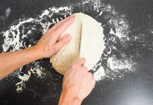 Person cutting a dough into two pieces on a black table with flour spread