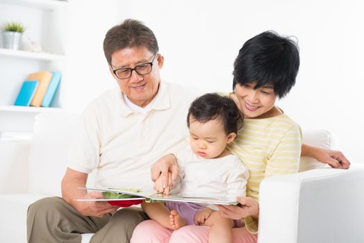 Asian family reading book sitting on sofa indoor, grandparents and grandchild living lifestyle at home.
