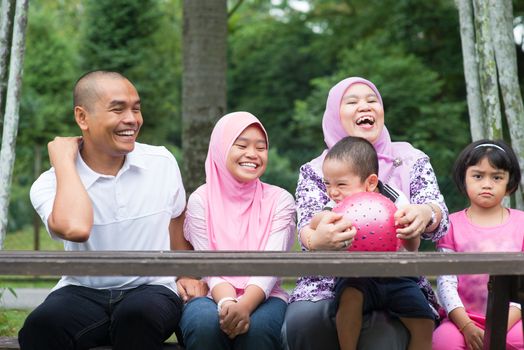 Happy Southeast Asian family sitting at garden bench laughing together, outdoor lifestyle at nature green park.