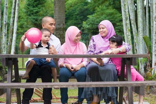 Happy Southeast Asian family sitting at garden bench chatting with each other, outdoor lifestyle at nature green park.