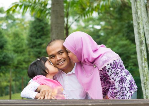 Happy Southeast Asian Muslim family, kissing on father face, outdoor lifestyle at nature green park.