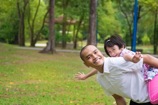 Father and daughter playing piggyback at outdoor garden park. Happy Southeast Asian family living lifestyle.