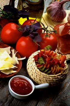 Arrangement of Raw Colorful Rotini Pasta and Ingredients with Grated Cheese, Smoked Parma Ham, Tomatoes, Spices, Olive Oil an Tomato Sauce closeup on Rustic Wooden background