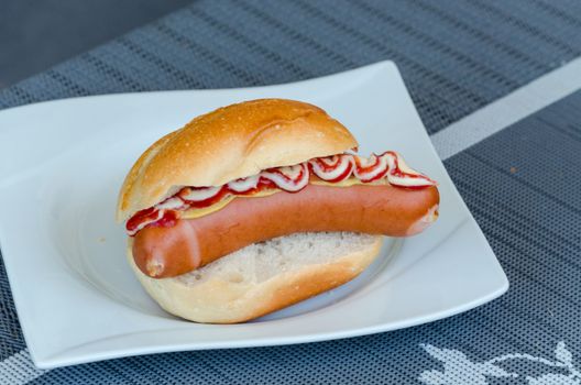 Hot dog with ketchup and mustard in bread roll on a white plate.