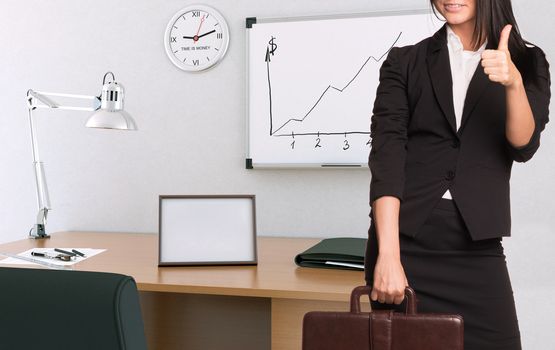 Businesswoman in suit holding briefcase and showing thumb-up. Office interior on background