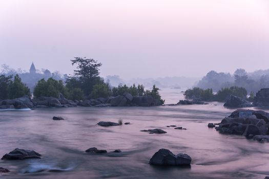 Morning fog at river flowing over dispersed rocks under pink skies reflected in the water.