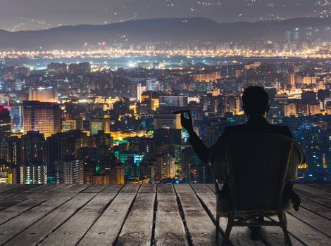 Silhouette of businessman sit on chair and hold a cigar and looking at the city in night.