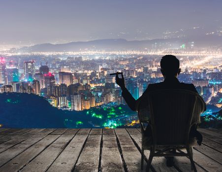 Silhouette of businessman sit on chair and hold a cigar and looking at the city in night.