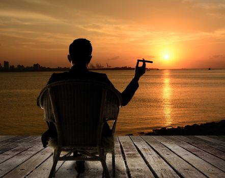 Silhouette of businessman sit on chair and hold a cigar and looking at the city in night.