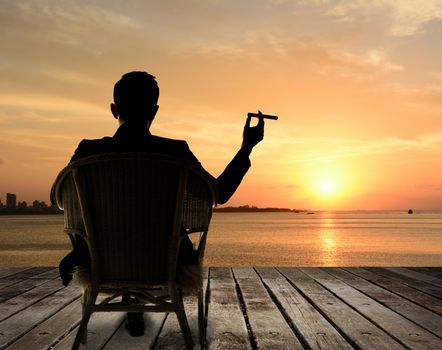 Silhouette of businessman sit on chair and hold a cigar and looking at the city in night.