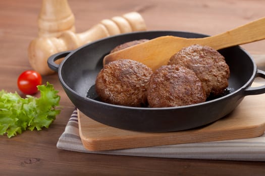 Meat rissoles in the iron pan with green lettuce and red tomato on the wooden table