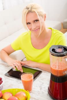 A beautiful mature woman with a smoothie in the kitchen using a tablet.
