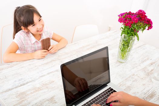 The daughter eating chocolate while her mother is working on a Laptop computer.