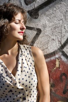A vintage style dressed girl leaning at a graffiti wall and enjoying the sunlight.
