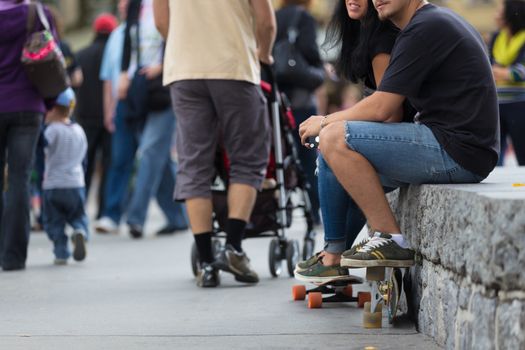 young couple with skateboards, sitting at the stone rail and waching people passing by. Urban scene.