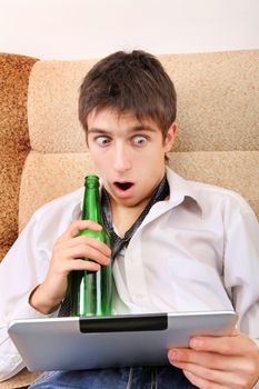 Surprised Teenager drinks a Beer with Tablet Computer on the Sofa at the Home