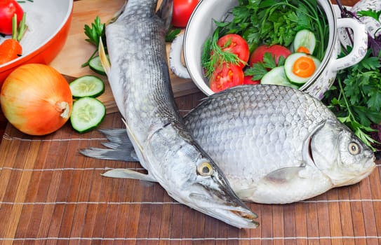 On a table on a chopping board there are a fish, cucumbers, tomatoes, onions, spices and parsley greens.