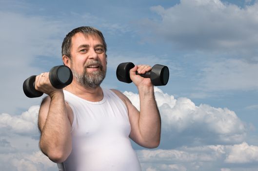 Elderly man exercising with dumbbells against cloudy sky background