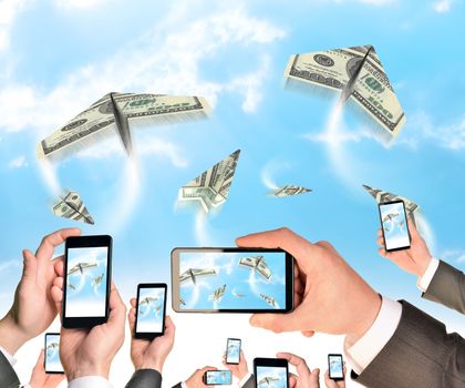 Hands holding smart phones and shoot video as flying paper plane of dollars. Sky with clouds on background