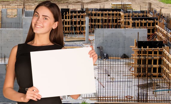 Beautiful woman smiling and hold empty paper sheet. Construction site as backdrop. Industrial concept