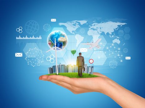 Hand holds city of skyscrapers on green grass and businessman walking forward. Earth and virtual elements near hand. Element of this image furnished by NASA