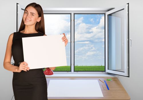 Beautiful businesswoman in dress smiling and holding empty paper sheet. Open window with green meadow and sky as backdrop