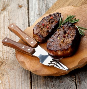 Roasted Beef Steaks with Spices and Rosemary on Wooden Plate with Fork and Table Knife on Rustic Wooden background