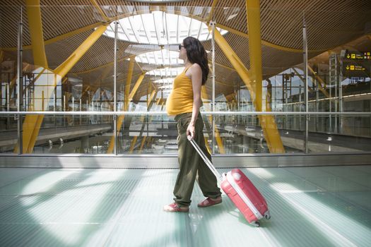 woman pregnant pulling suitcase inside airport hall