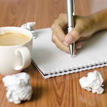 woman hand writing with pen on notebook.there are crumpled paper and coffee cup on wood table background