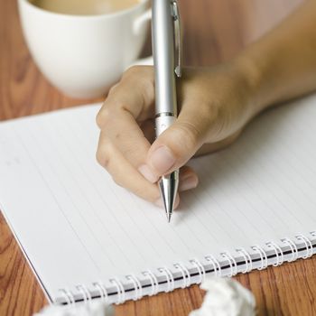 woman hand writing with pen on notebook.there are crumpled paper and coffee cup on wood table background