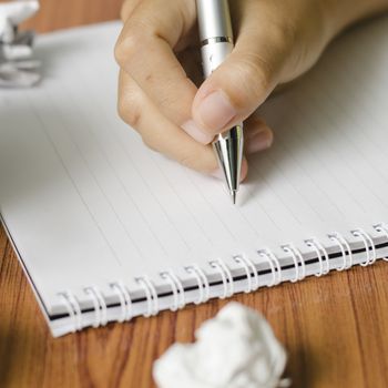 woman hand writing with pen on notebook.there are crumpled paper and coffee cup on wood table background