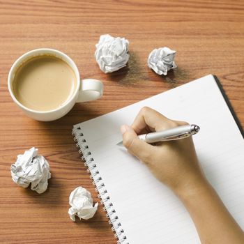 woman hand writing with pen on notebook.there are crumpled paper and coffee cup on wood table background