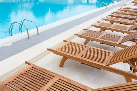 modern wooden sunbeds by turquoise swimming pool. 
