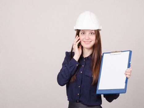 Young woman builder in helmet standing with a folder in hand and talking on phone