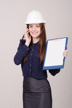 Young woman builder in helmet standing with a folder in hand and talking on phone