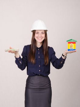 Young woman builder in helmet with little house and worth the money in the hands of