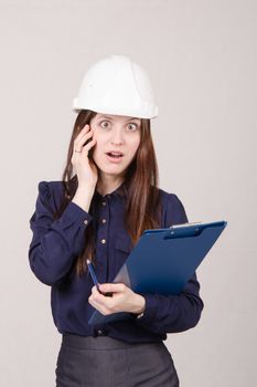 Young woman builder in helmet standing with a folder in hand and talking on phone