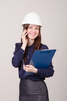Young woman builder in helmet standing with a folder in hand and talking on phone