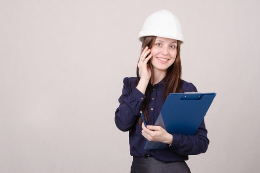 Young woman builder in helmet standing with a folder in hand and talking on phone